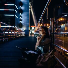 side view of woman listening music in headphones with laptop on knees on city street at night