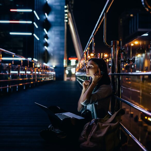 side view of woman listening music in headphones with laptop on knees on city street at night